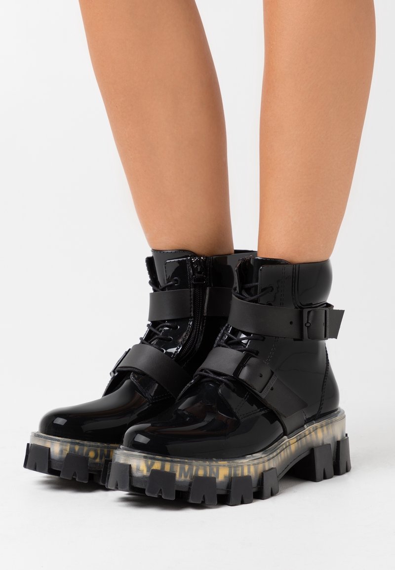 Bottes de cheville en cuir verni noir avec semelle épaisse et dentelée, sangles réglables et fermeture éclair latérale. Présente une plateforme translucide avec des accents de texte.
