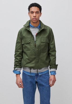 Junger Mann trägt eine grüne Jacke über einem grauen Sweatshirt und einem blauen Jeanshemd, kombiniert mit blauen Jeans, steht vor einem schlichten Hintergrund.