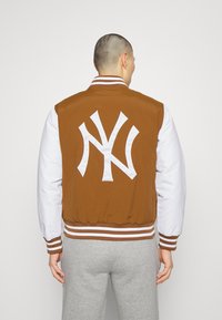 Brun och vit varsityjacka med en framträdande vit logotyp på ryggen, randiga ribbade muddar och nederkant, med en slät textur.