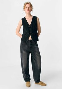 Schwarze Denim-Weste mit V-Ausschnitt und zwei vorderen Knöpfen, kombiniert mit locker sitzenden schwarzen Jeans und beigen Slippern. Minimalistisches Design.