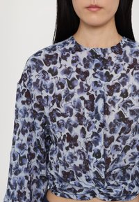 Blouse fleurie aux nuances de bleu et de violet sur un fond clair. Coupe ample avec manches longues et détail noué à l'ourlet.