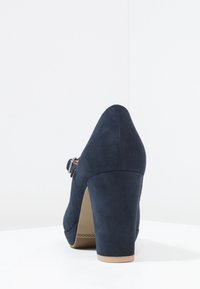 Escarpin en suede bleu marine avec un bout arrondi, un talon carré de hauteur moyenne et une bride de cheville avec un détail de boucle en argent.