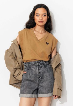 Jeune femme portant un top beige sans manches avec un détail en forme de cœur, un short en jean gris taille haute roulé et une veste beige drapée sur ses épaules.