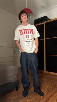 Biele tričko s červeným grafickým motívom "NIKE 58", voľné modré džínsy a čierne topánky. Izba s drevenou podlahou a tmavým nábytkom v pozadí.