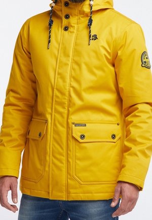 Homme portant une veste à capuche jaune vif avec des poches avant et des détails de boutons noirs, associée à un jean bleu.