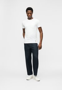 Νέος άνδρας που στέκεται μπροστά σε λευκό φόντο, φορώντας λευκό κοντομάνικο πουκάμισο, σκούρα μπλε τζιν και λευκά αθλητικά παπούτσια.