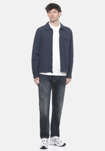 Uomo in piedi con indosso una giacca a zip blu navy, una t-shirt bianca, jeans scuri e sneakers bianche su uno sfondo chiaro e semplice.
