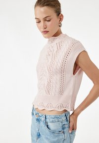Jasnoróżowy, dziergany crop top z koronkowymi wzorami i wysokim kołnierzem. Posiada krótkie rękawy i falbaniaste wykończenie. Połączony z jasnoniebieskimi dżinsami.