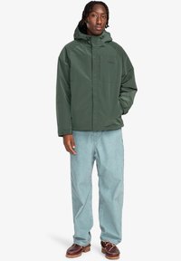 Green, waterproof jacket with a hood, front pocket, and adjustable drawstrings, paired with light blue loose-fit pants and brown shoes.