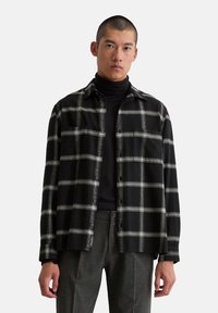 Chemise à carreaux noir et blanc avec un devant boutonné, deux poches poitrine et une coupe décontractée, superposée à un turtleneck noir.