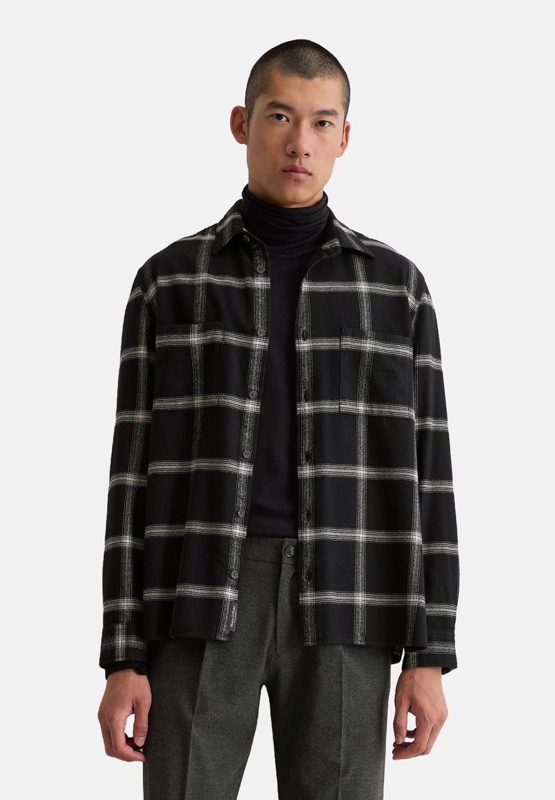Chemise à carreaux noir et blanc avec un devant boutonné, deux poches poitrine et une coupe décontractée, superposée à un turtleneck noir.