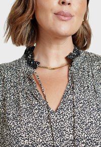 Blusa morbida con stampa leopardata nera e crema, caratterizzata da un collo scallop e da pieghe leggere sulle spalle. Collana in tonalità oro visibile.