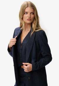 Navyblauwe pinstripe blazer met een sluiting met één knoop en zijzakken, gedragen over een bijpassend button-up top met een V-hals.