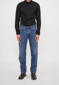 Blå denimjeans med rak passform, fem fickor och knappstängning, i kombination med en svart långärmad skjorta och svarta stövlar.