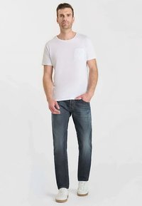 T-shirt blanc à manches courtes avec une poche poitrine, associé à un jean slim bleu foncé et des baskets blanches à semelles beige. Texture du tissu lisse.