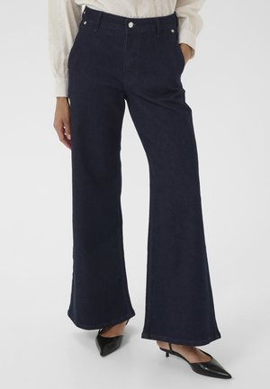 Een persoon draagt donkere wijde jeans in combinatie met een lichte, gestructureerde blouse en zwarte, puntige schoenen, staande tegen een effen witte achtergrond.