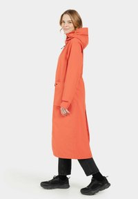 Oranje lange waterdichte trenchcoat met een capuchon, verstelbare taille, zijzakken en een rechte pasvorm, gecombineerd met zwarte sneakers en broek.