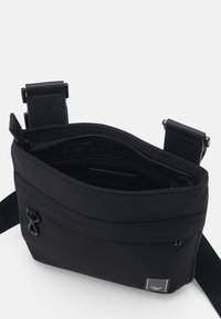 Svart axelremsväska med en mjuk nylonutsida, fack med dragkedja och justerbar rem, med en liten logotyp och en slät textur.