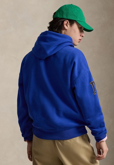 Jeune personne portant un sweat à capuche bleu vif et un pantalon kaki, coiffée d'une casquette de baseball verte, posant devant un fond beige neutre.