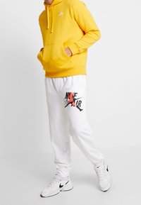 Sudadera con capucha amarilla y bolsillo frontal, combinada con pantalones de chándal blancos que presentan un gráfico en negro y rojo de 'Nike Air' y zapatillas a juego.