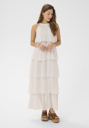 Vrouw met lang haar die een mouwloze witte jurk met meerdere lagen en zwarte stippen draagt, gecombineerd met bruine platform sandalen, terwijl ze voor een effen achtergrond staat.