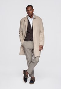 Abrigo beige tipo trench superpuesto sobre un cárdigan marrón, combinado con pantalones gris claro y zapatos oscuros con cordones marrones. Diseño simple y limpio.