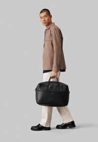 Sac de sport en cuir noir avec double poignées et fermeture éclair, présenté par une personne portant une veste beige et un pantalon blanc cassé.