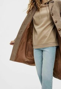 Brun hundtandsmönstrad kappa med fodrad insida, bärs över en beige hoodie och ljusblå jeans. Har knapprade detaljer och en avslappnad passform.