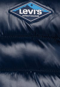 Navyblauwe pufferjack met een glanzende afwerking en zichtbare naden; heeft een diamanten Levi's-logo in wit en blauw op de borst.