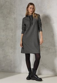 Mekko, jossa on pitkät hihat, musta ja harmaa väri, kolmiomainen kuvio, nyörikiinnitteinen huppu ja sivurei'ät, yhdistettynä mustiin nilkkabootseihin.