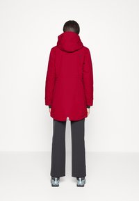 Chaqueta impermeable roja con capucha, puños elásticos y espalda ventilada. Combinada con pantalones negros y zapatos grises con acentos en azul.