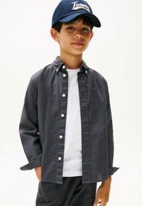 Camisa de botones gris con una camiseta blanca debajo, combinada con pantalones oscuros. Una gorra azul con el logo de "Tommy" al frente. Diseño casual.