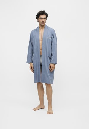 Homme debout pieds nus portant un peignoir bleu clair longueur genoux par-dessus des sous-vêtements gris sur fond blanc uni