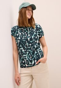 Mujer con una gorra azul verdosa, camisa con un patrón abstracto negro y azul verdosa, y pantalones beige, apoyada contra una pared de color claro, mirando a la derecha.