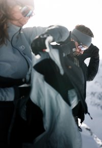 Two individuals in outdoor gear. One wears a light gray jacket, black gloves, and mirrored sunglasses, while the other dons a black jacket and gray headband.