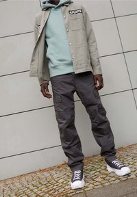 Grau leichte Jacke über einem mintgrünen Hoodie, kombiniert mit dunklen Cargo-Hosen und schwarz-weißen Sneakers, was einen geschichteten Look kreiert.