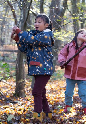 Two children outdoors in autumn. One wears a navy jacket with orange and yellow celestial prints, burgundy pants, and yellow boots. The other wears a pink jacket.