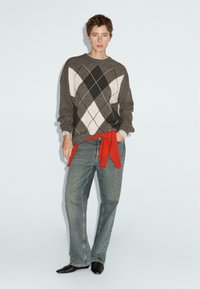 Giovane in jeans larghi blu, maglione argyle marrone, felpa rossa legata in vita e scarpe nere a punta, contro uno sfondo neutro.