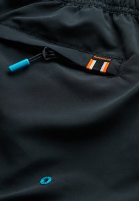 Pantaloni tecnici neri con coulisse blu, etichetta a strisce arancioni e bianche, e un'etichetta con un emblema turchese.
