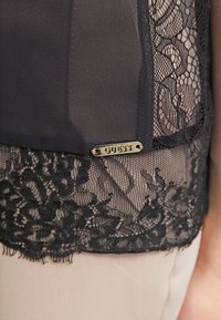 Sort top med blonde detaljer i kanten, samt et guld "GUESS" logo mærke. Glat stof, indviklede blonde mønstre og en flatterende pasform.