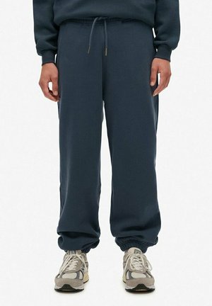 Person iført mørk marineblå joggingbukser med snøre i taljen og elastiske manchetter, kombineret med beige og grå sneakers.