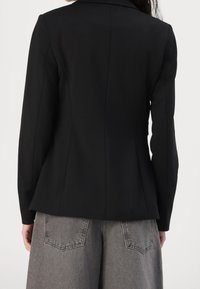 Femme portant un blazer noir ajusté et un jean gris ample, vue de dos, debout devant un fond blanc uni.