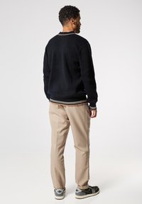 Maglione nero lavorato a maglia con texture a costine e bordo bianco e beige nella parte inferiore, abbinato a pantaloni beige e sneaker nere e grigie.