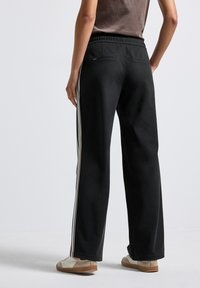 Personne portant un pantalon large noir avec des bandes blanches sur les côtés et des baskets beiges, se tenant devant un fond clair uniforme.