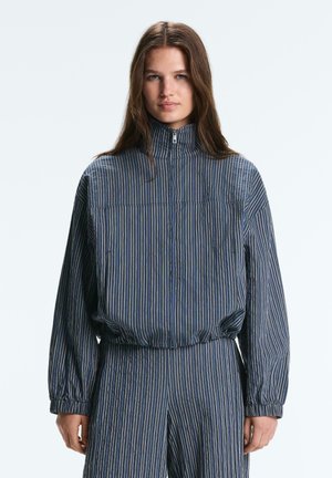 Frau steht, trägt eine blau gestreifte Zip-up-Jacke und passende gestreifte Hose, mit langen braunen Haaren und neutralem Gesichtsausdruck.