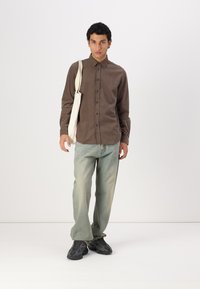 Braunes Hemd mit Knöpfen, helle Jeans, schwarze Sneaker mit strukturiertem Muster, hält eine weiße Tragetasche. Sauberer, minimalistischer Hintergrund.