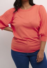 Femme portant un haut en maille corail à manches volumineuses et un jean en denim bleu, posant avec une main sur la hanche devant un fond uni.