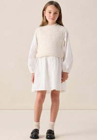 Weißes Baumwollkleid mit einem strukturierten Strick-Oberteil ohne Ärmel. Die Bluse hat puffige Ärmel, und das Outfit wird mit schwarzen Schuhen und beigen Socken kombiniert.