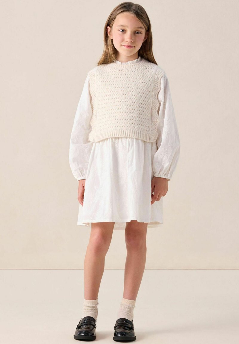 Weißes Baumwollkleid mit einem strukturierten Strick-Oberteil ohne Ärmel. Die Bluse hat puffige Ärmel, und das Outfit wird mit schwarzen Schuhen und beigen Socken kombiniert.