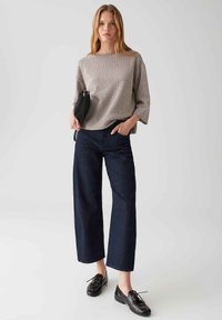 Jeune femme debout, portant un haut ample beige à motifs, un jean large noir, des chaussures en cuir noires et tenant un sac à main noir.